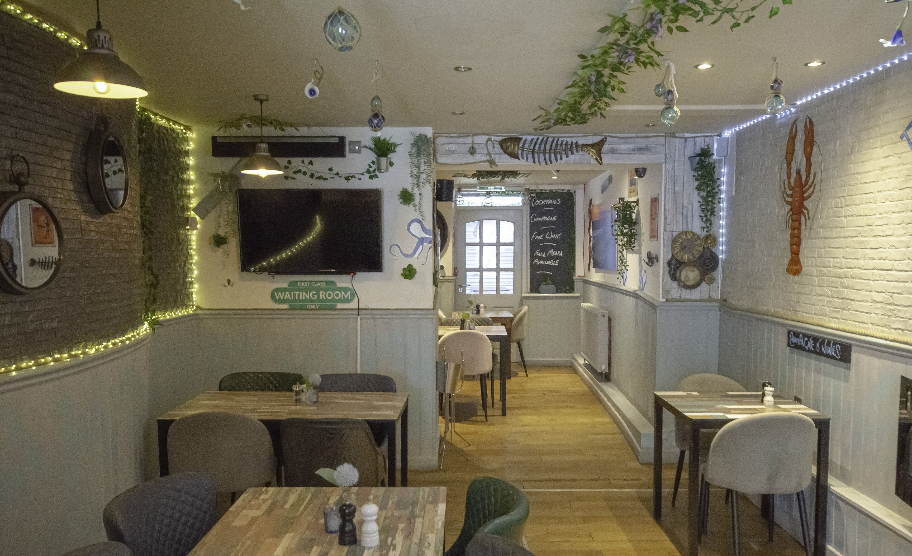 Interior d'un restaurant amb paret de maó, plantes penjants i un menú escrit a la pissarra. Taules i caires disposades per dinar.