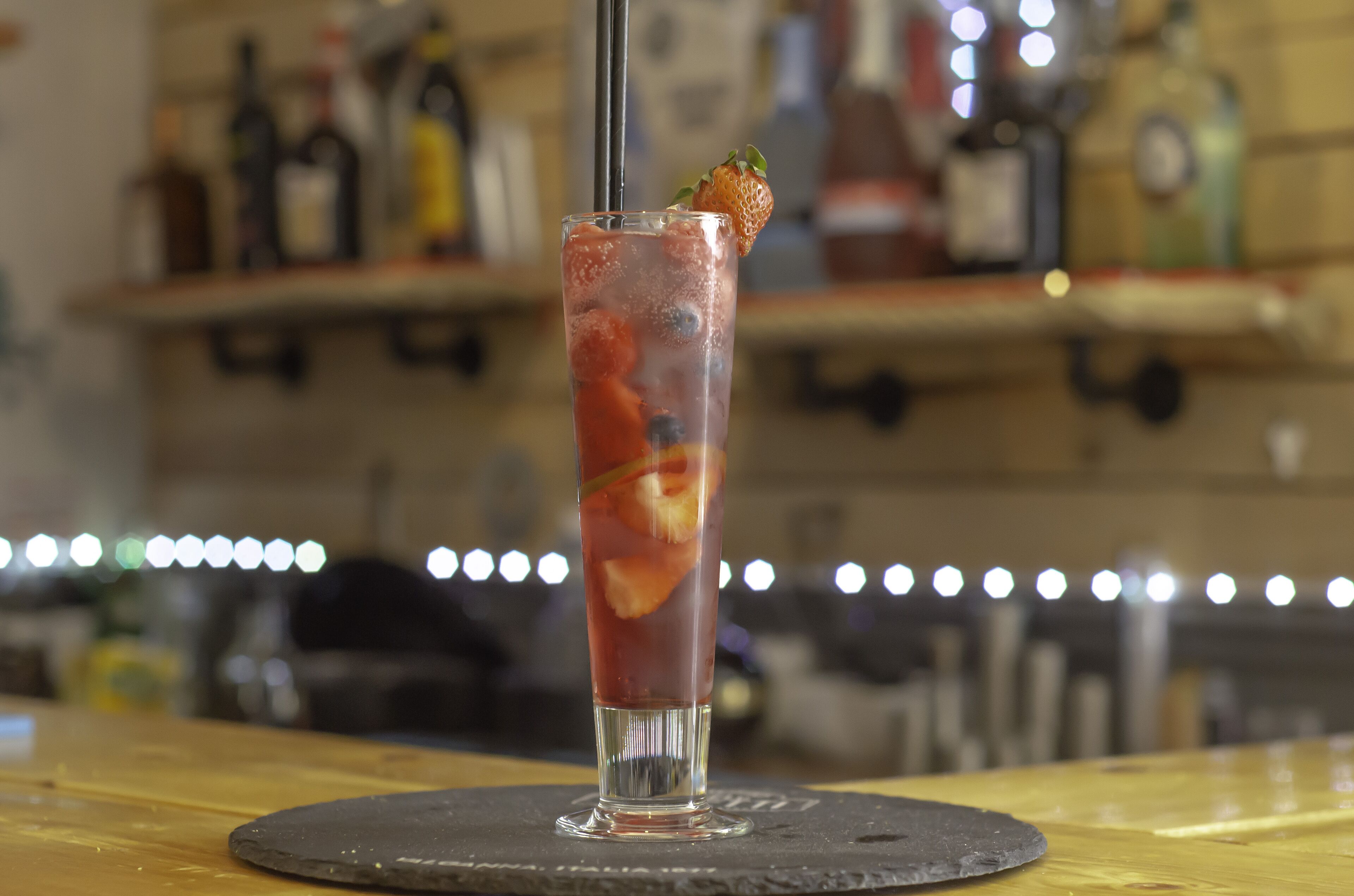 A tall glass with a strawberry garnish, filled with a red beverage and ice cubes, placed on a coaster.