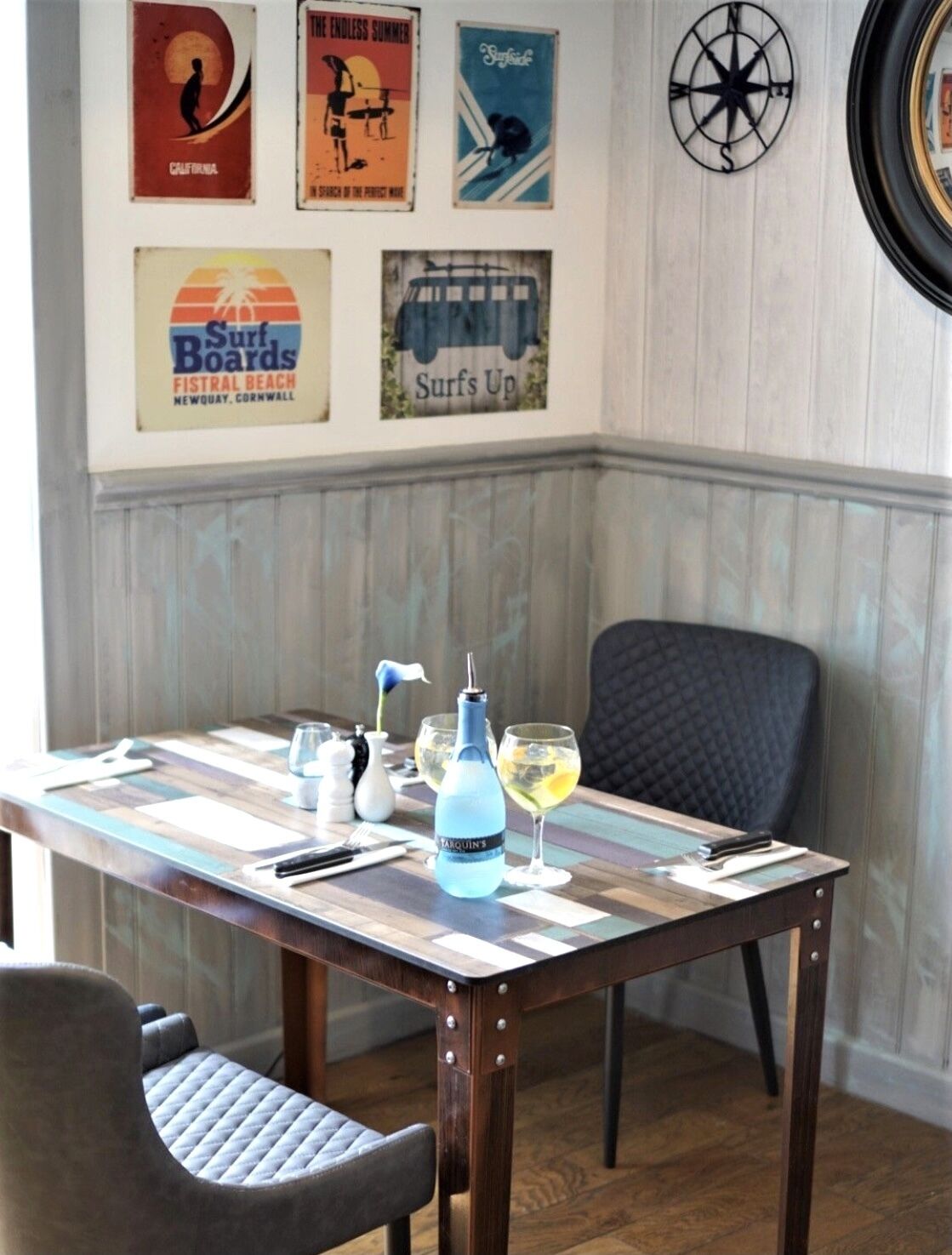 Una mesa de comedor con una botella azul, copas de vino y cubiertos, frente a una pared decorada con carteles de surf antiguos.