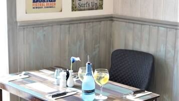 Una mesa de comedor con una botella azul, copas de vino y cubiertos, frente a una pared decorada con carteles de surf antiguos.