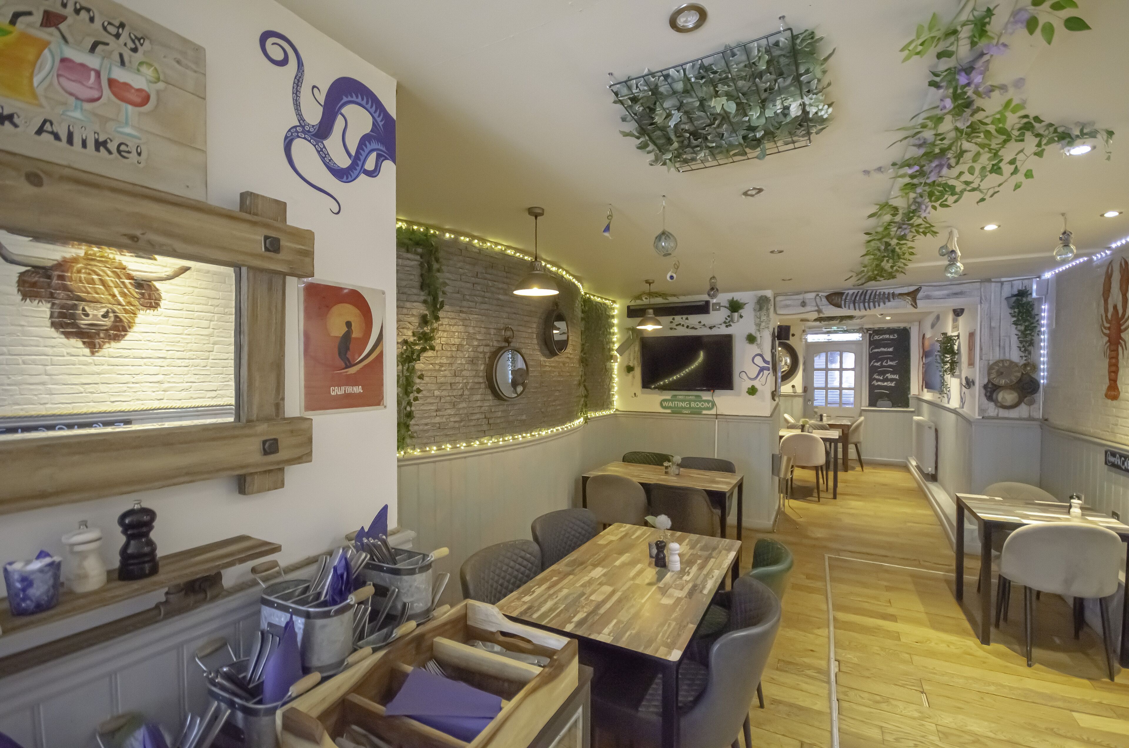A restaurant interior with wooden tables, chairs, a TV mounted on the wall, and decorative plants.