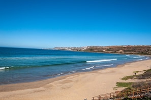 A beach with a wooden walkway leading to the shore, a clear blue sky, and a distant view of a town.