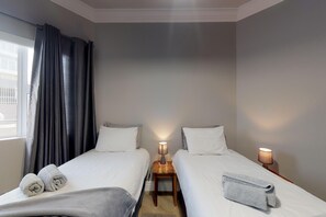 A hotel room with two beds, each with a pillow and a rolled towel. There are two bedside tables with lamps.