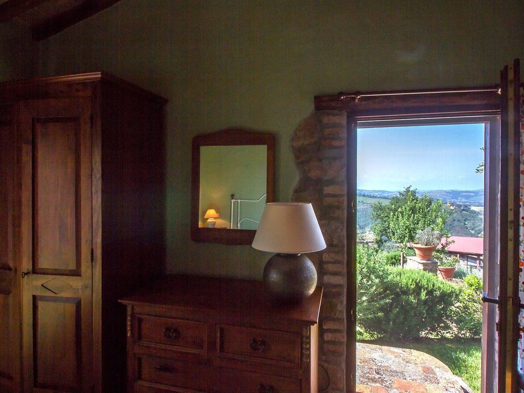 Une pièce avec une commode en bois, une lampe et un miroir. De la fenêtre, on aperçoit des arbres et des bâtiments.
