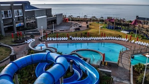 Un espace piscine d’un complexe hôtelier comprenant un grand toboggan aquatique, des chaises longues et une vue sur la mer.