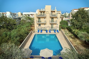 A swimming pool with blue lounge chairs and umbrellas, surrounded by greenery and a building with balconies. - Acropolis Apartments (Hersonissos)