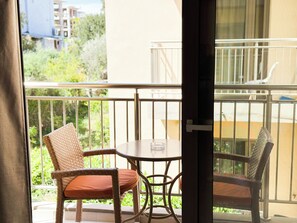 Un balcon avec des fauteuils en osier et une table, une vue sur la verdure et les bâtiments, et une porte partiellement visible.