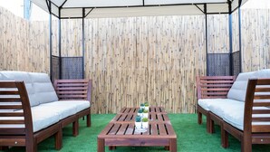 A covered patio area with two wooden sofas, a wooden table, and a bamboo fence. - Salame TLV Apartments (Tel Aviv)