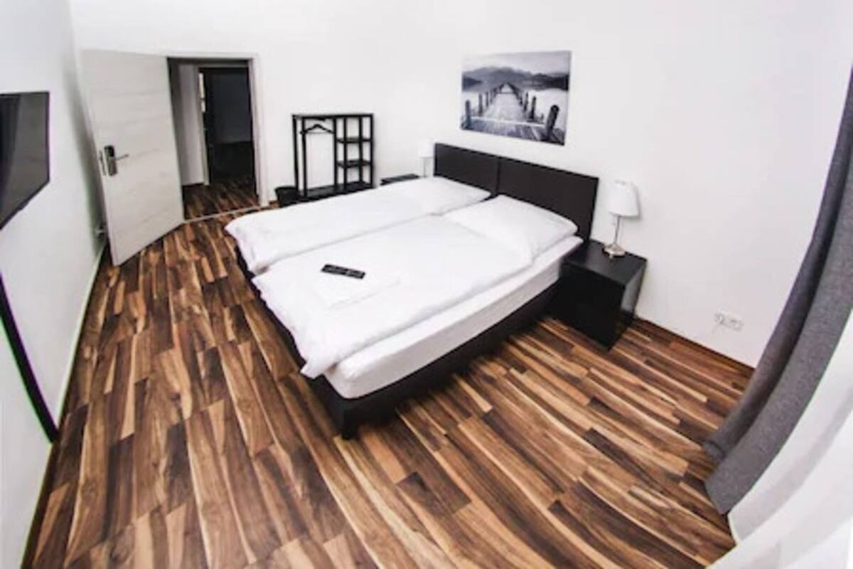 A bedroom with a wooden floor, a bed, a nightstand, a lamp, and a picture on the wall.