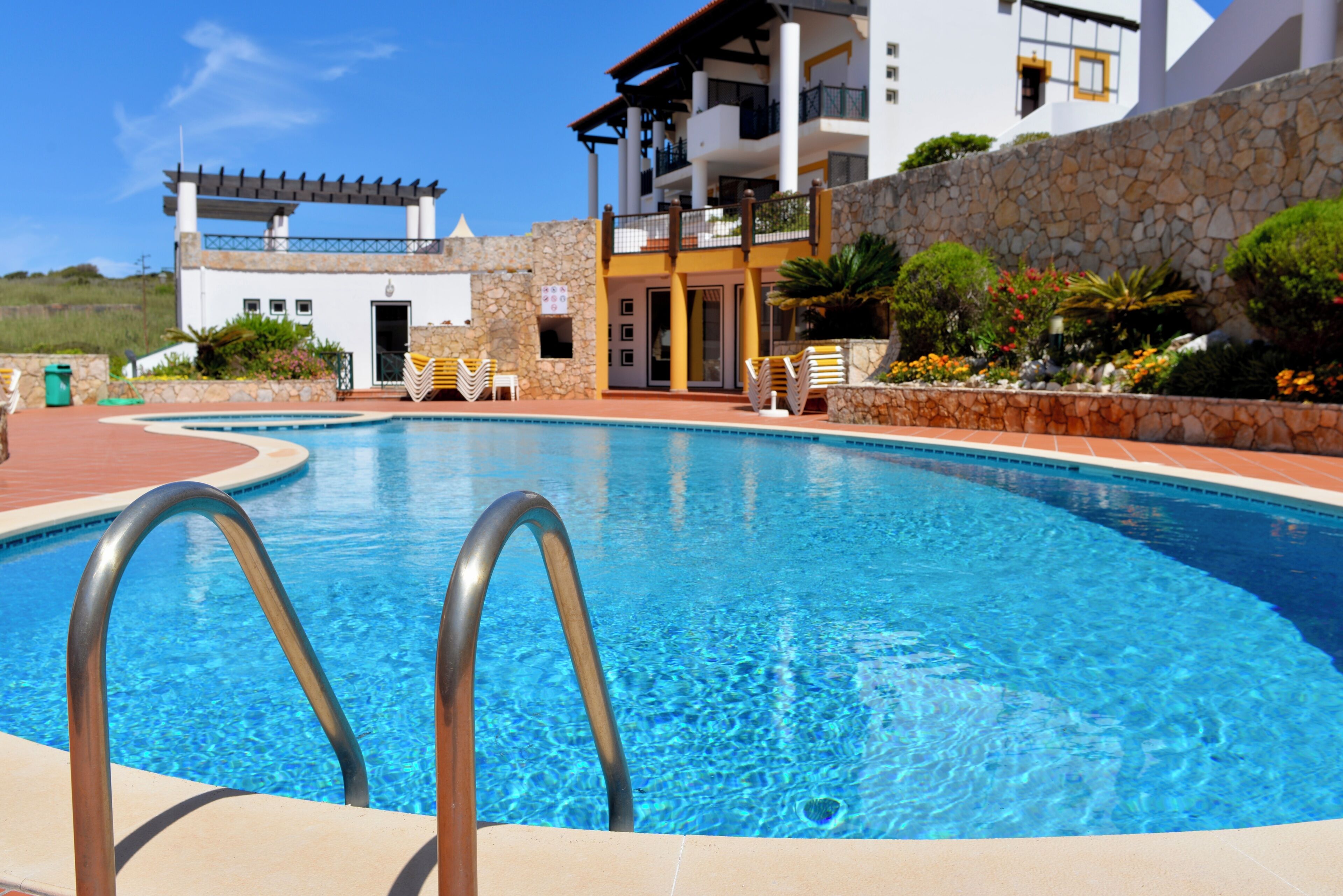 A swimming pool with metal ladders, surrounded by a stone wall and a building with balconies.