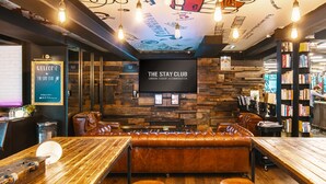 Un espace salon confortable et bien éclairé, avec une table en bois, des sièges en cuir et un grand écran de télévision diffusant des images de « The Stay Club London Student Accommodation ».