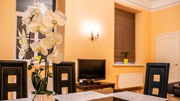 Un coin repas avec une table dressée pour quatre personnes, un vase rempli d’orchidées blanches, un téléviseur et une fenêtre avec des stores.