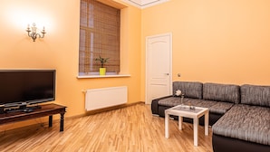 Un salon avec un téléviseur posé sur un meuble en bois, un canapé, une petite table et une fenêtre avec des stores.