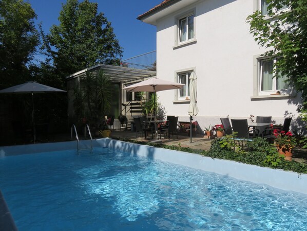 A swimming pool with a sunshade, outdoor seating, and potted plants. - Pension Säntisblick (Meersburg)