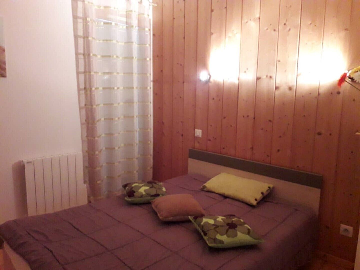A bedroom with a wooden wall, a bed with purple bedding, and a window with a sheer curtain.