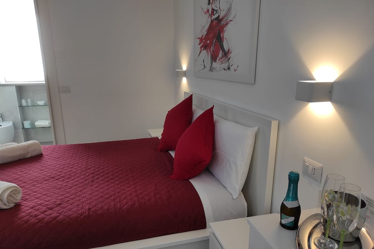 A hotel room with a red bedspread, white pillows, a bedside table with a bottle and glasses, and a wall-mounted light.