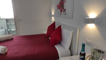 Una habitación de hotel con una colcha roja, almohadas blancas, una mesita de noche con una botella y vasos, y una lámpara fijada a la pared.
