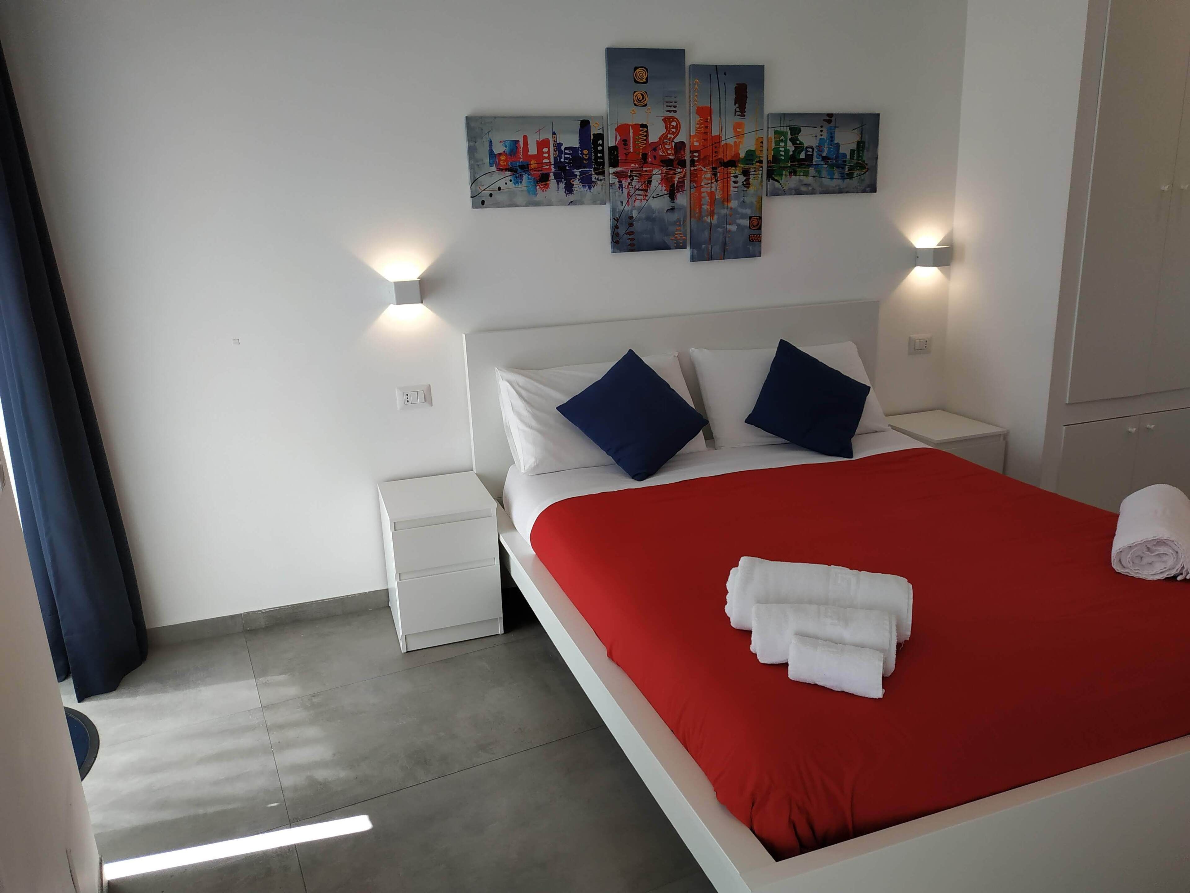 A hotel room with a bed, two nightstands, a red bedspread, white pillows, and a wall art piece above the bed.