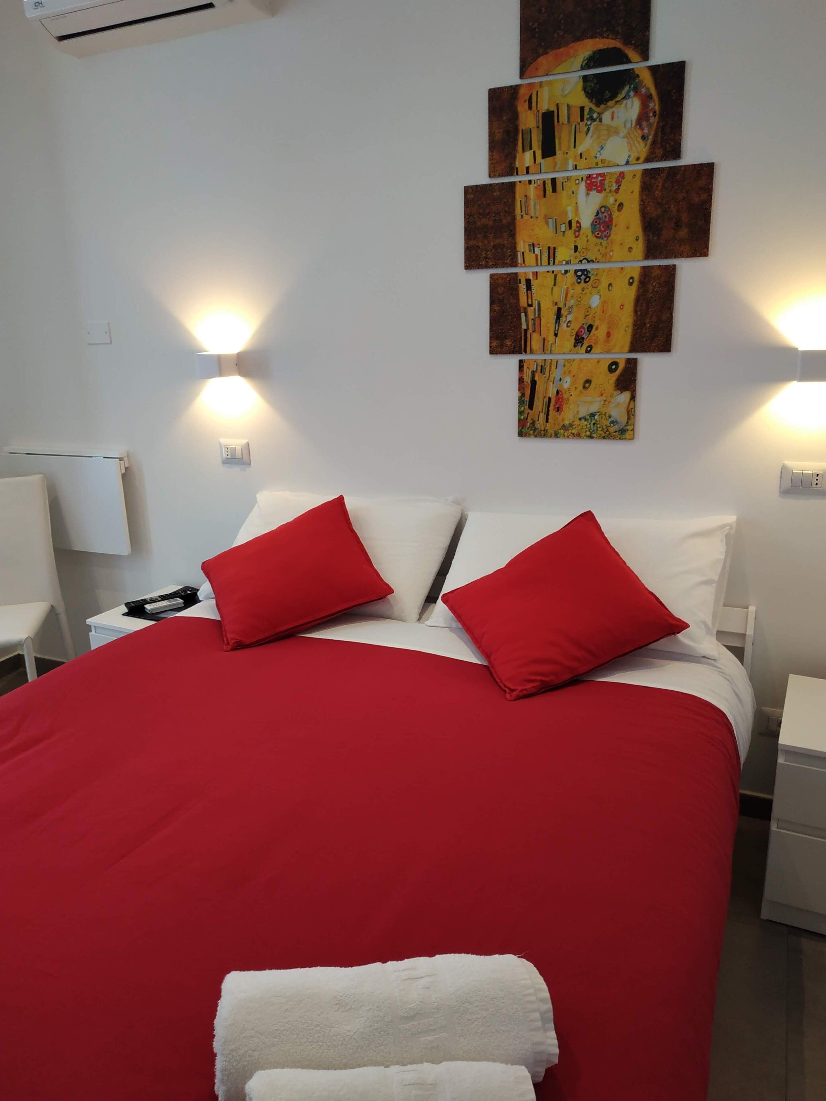 Habitación de hotel con una cama grande, ropa de cama roja y blanca, un cuadro en la pared y lámparas de noche.