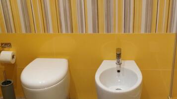Une salle de bain avec des toilettes et un bidet, des murs jaunes, un papier peint à rayures et une poubelle.