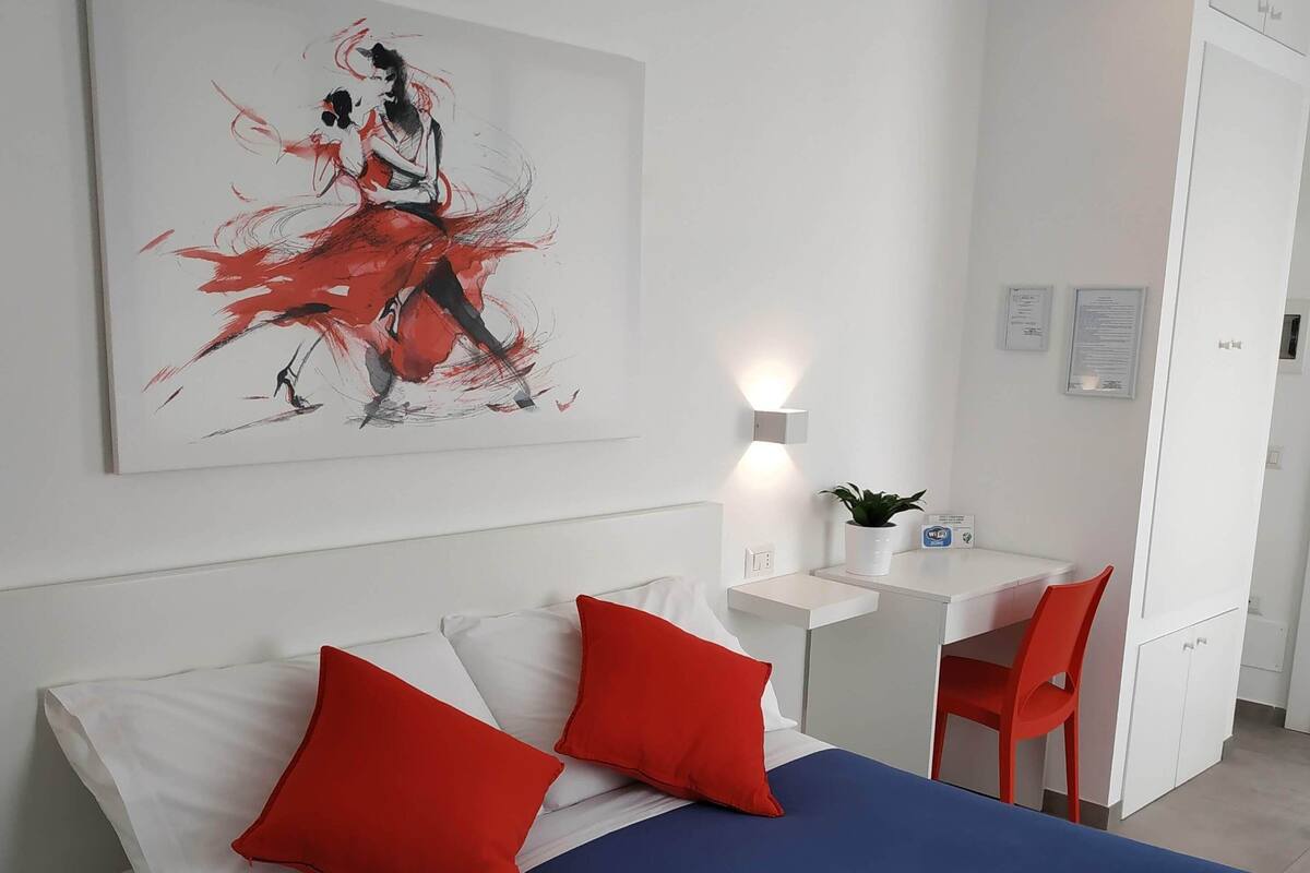 A modern hotel room with a large bed, red pillows, a desk, and a chair.