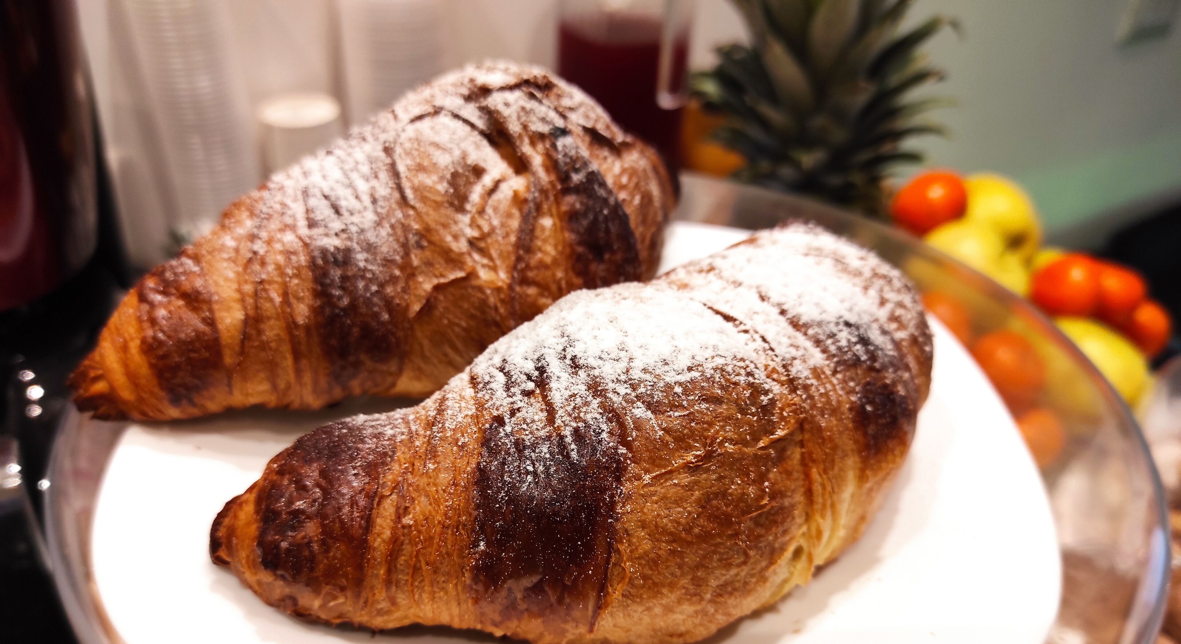Two croissants on a plate with a fruit bowl in the background.