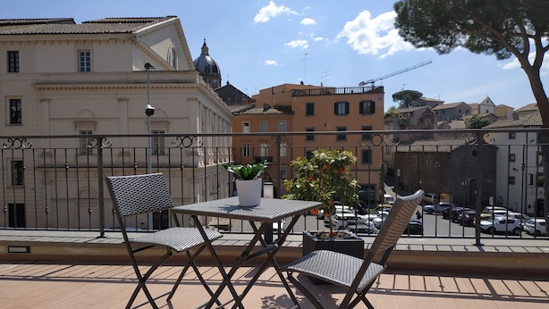 A terrace with a table and two chairs, overlooking a street with parked cars and buildings. - Palazzo Verdi B&B (Viterbo)