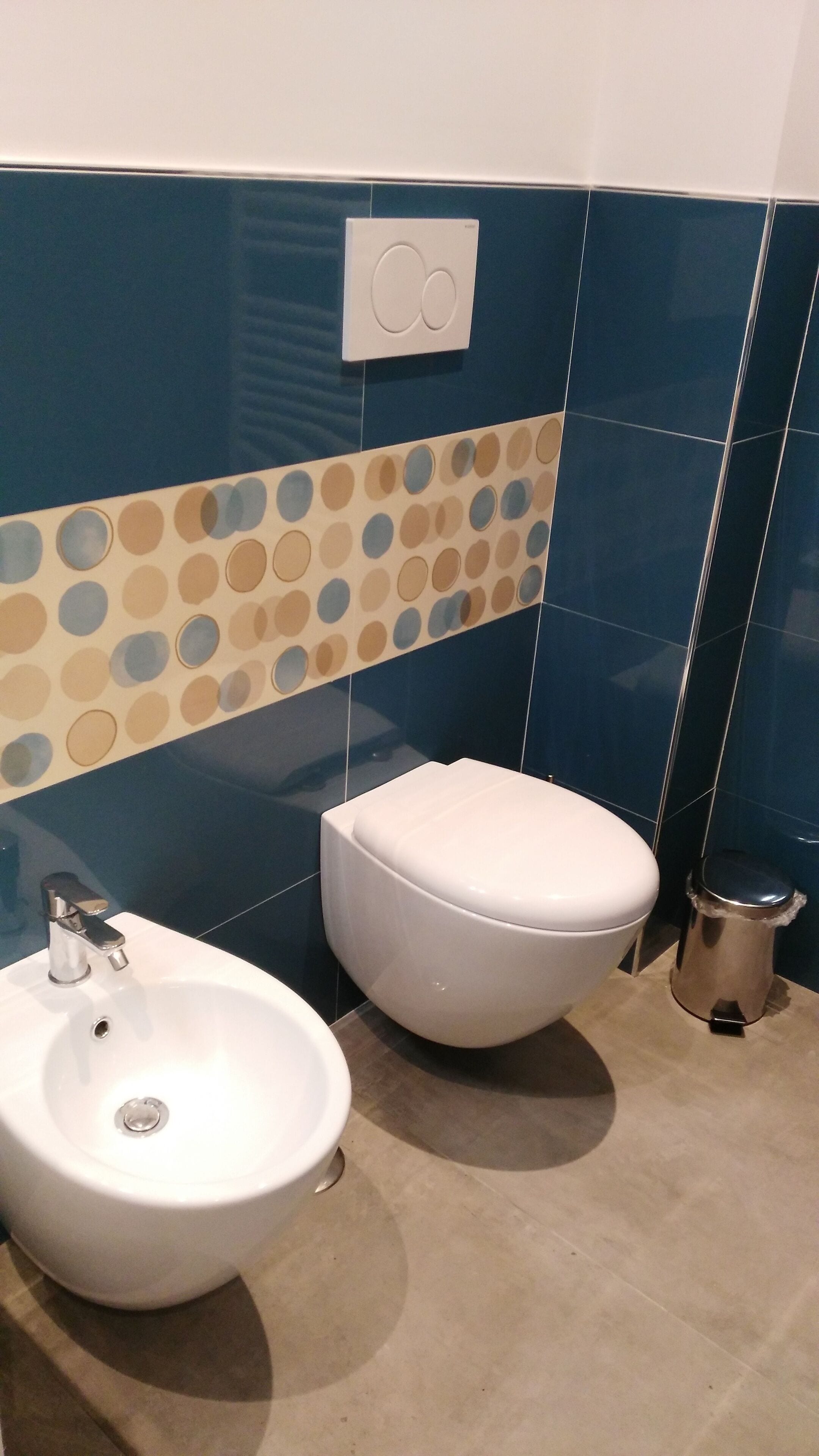 Ein Badezimmer mit weißem Waschbecken, WC und Bidet. Die Wände sind in Blau- und Beigetönen mit einem Punkte-Muster gefliest.