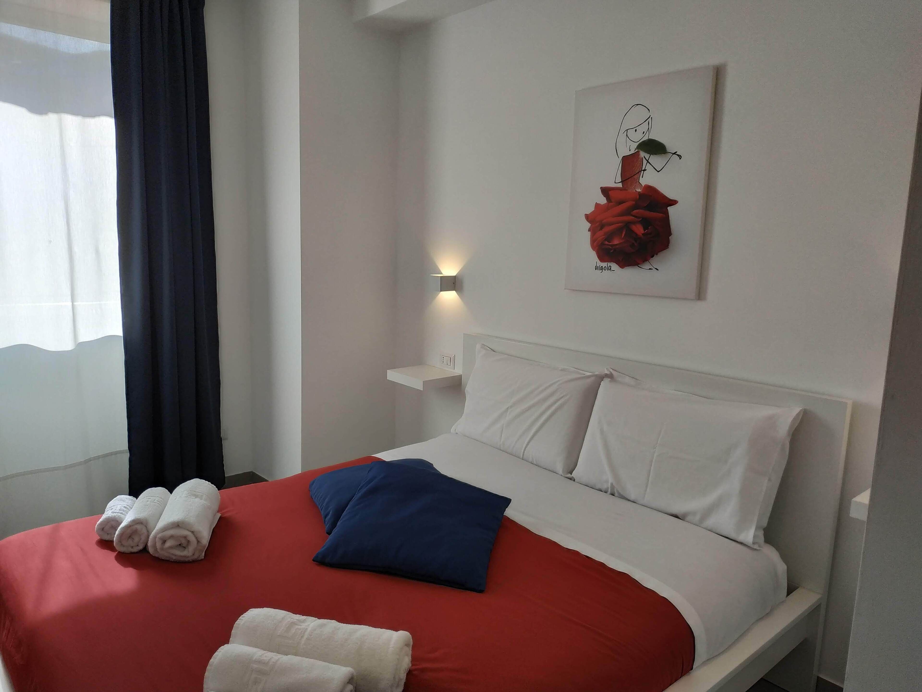 A hotel room with a bed, white pillows, a red blanket, and a wall-mounted artwork of a rose.