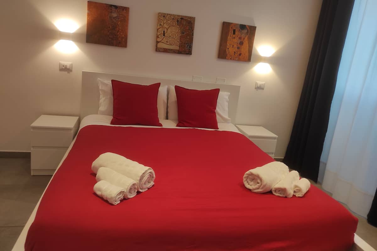A hotel room with a large bed, red bedding, two red pillows, white nightstands, and three wall-mounted paintings.