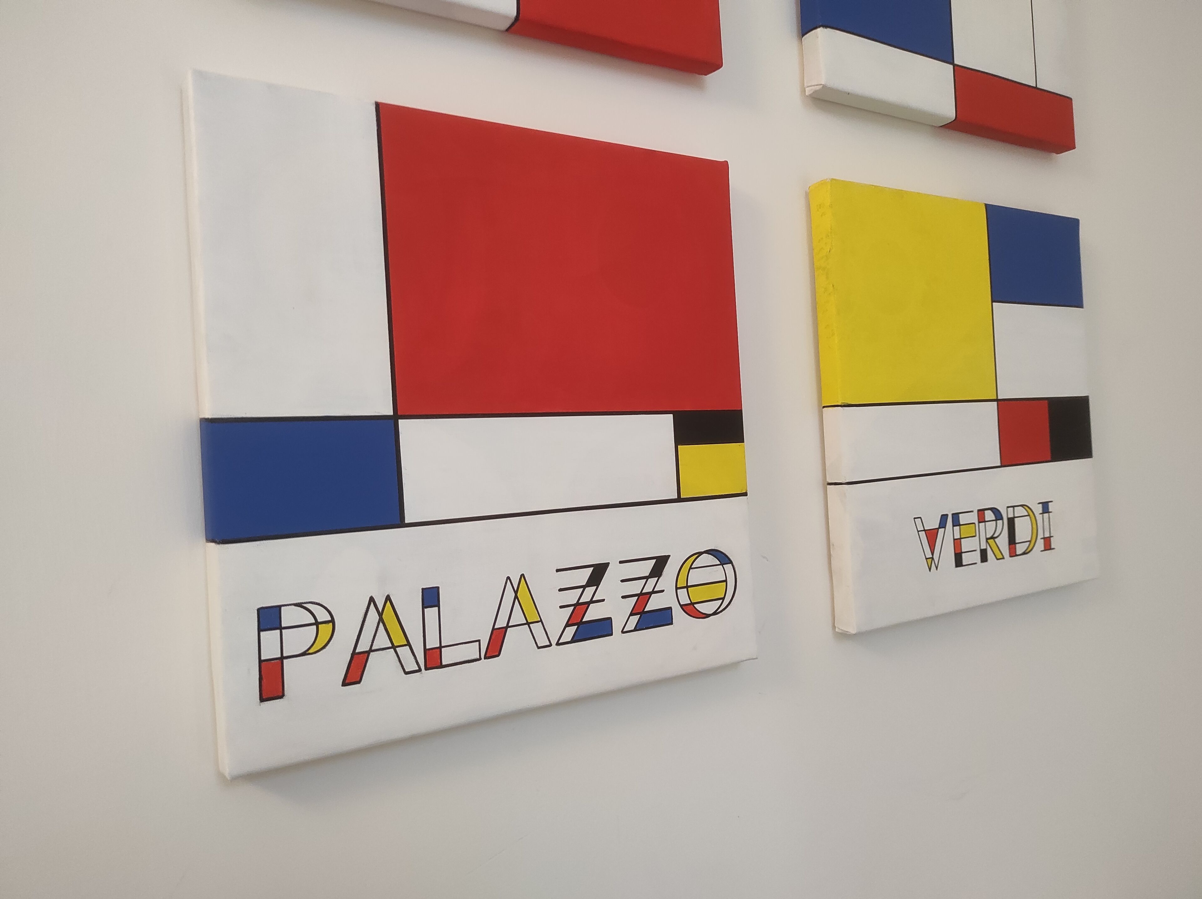 Two artworks with geometric shapes and bold colors, titled 'PALAZZO' and 'VERDI', on a plain wall.