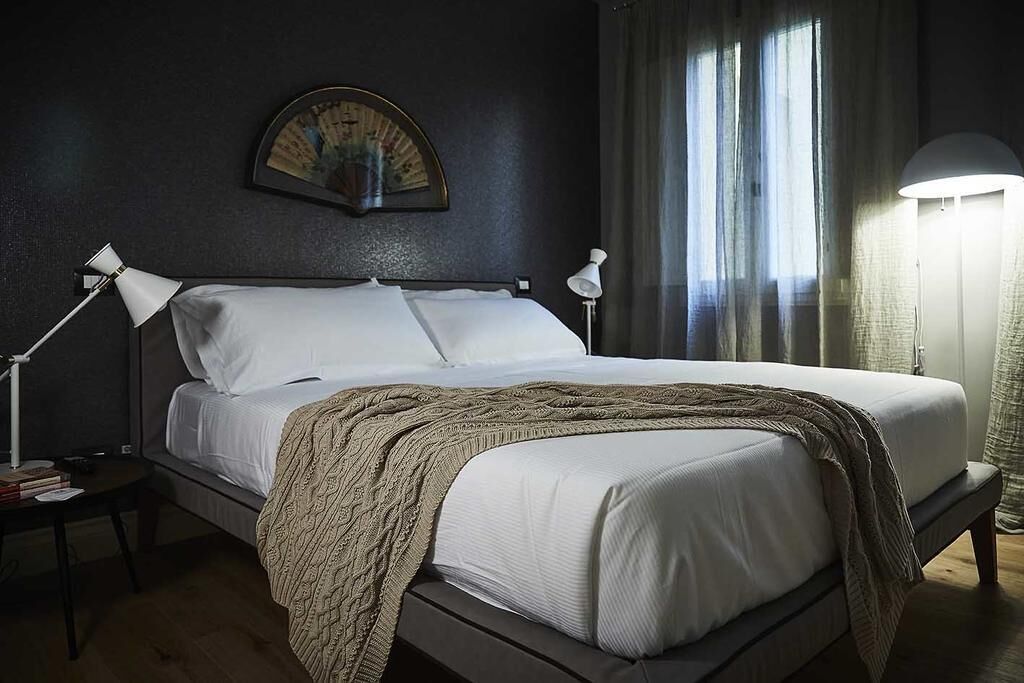 Una cama impecablemente tendida con sábanas blancas y una manta marrón, flanqueada por dos lámparas de noche sobre una mesita de noche de madera oscura.
