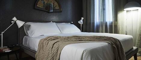 Una cama impecablemente tendida con sábanas blancas y una manta marrón, flanqueada por dos lámparas de noche sobre una mesita de noche de madera oscura.