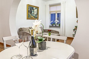 A modern living room with a white sofa, a dining table with three wine glasses and a bottle, a painting on the wall, and a window with curtains.