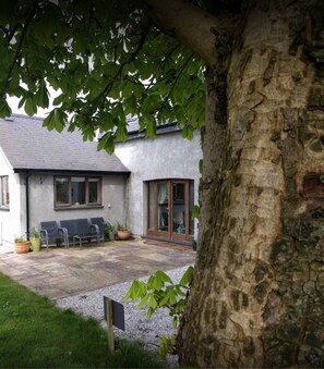 A house with a stone exterior, a patio with seating, and a tree in the foreground. - Honey Cottage (Amlwch)