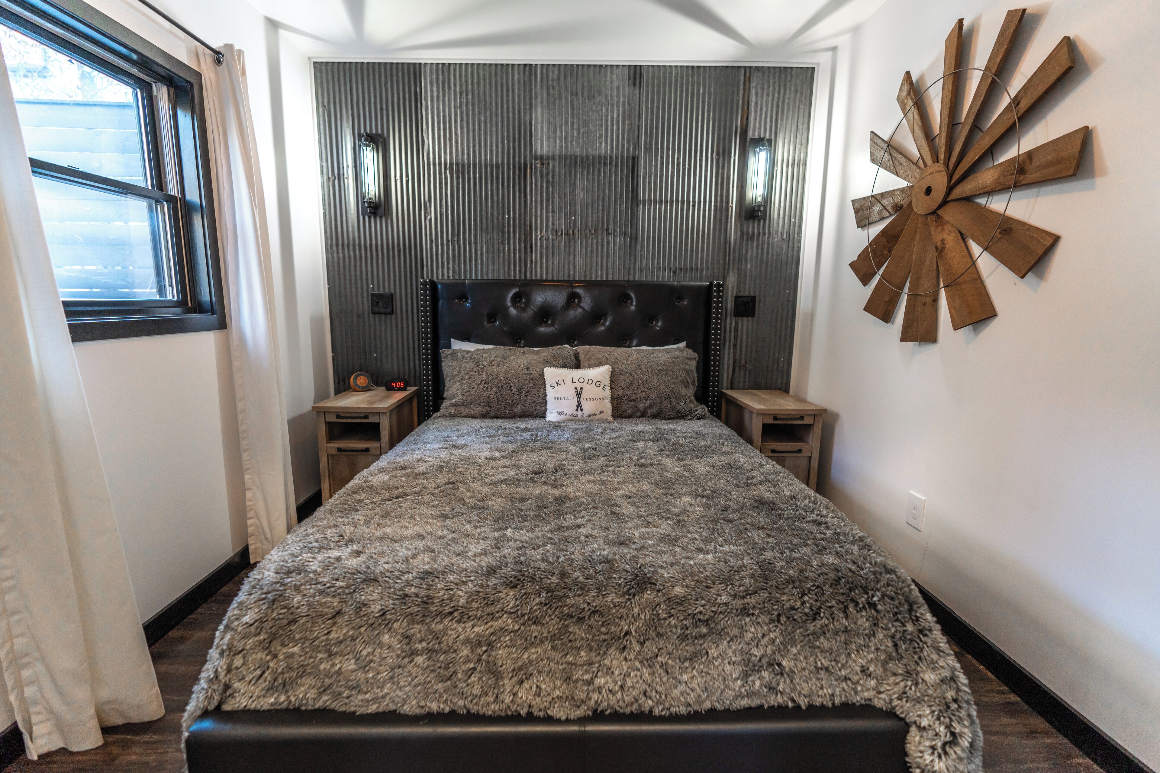 A bedroom with a tufted headboard, a fluffy gray bedspread, two bedside tables, and a wooden wall decor piece.
