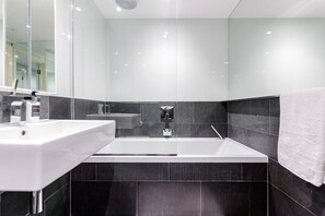 A modern bathroom with a white sink, a large bathtub, and dark tiled walls.