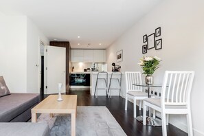 A modern living room with a sofa, a coffee table, and a dining area with white chairs and a glass table. A kitchen is visible in the background.
