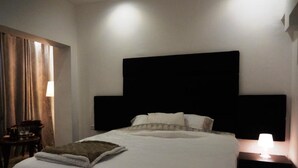 A neatly made bed with a white comforter and a folded blanket, a dark headboard, a bedside table with a lamp, and a chair with a table and a glass. - Gorgeous Studio On Hilton Beach (Tel Aviv)