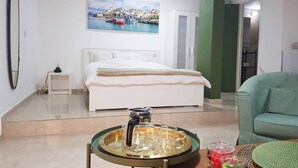 A hotel room with a bed, a painting of a coastal town, a green armchair, and a table with a glass pitcher and cups. - Large Beautiful Studio in Old Jaffa (Tel Aviv)