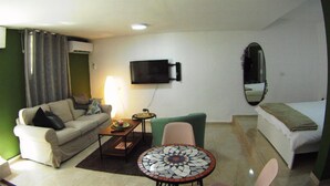 A compact living space with a sofa, a small table, a TV mounted on the wall, and a bed. - Large Beautiful Studio in Old Jaffa (Tel Aviv)