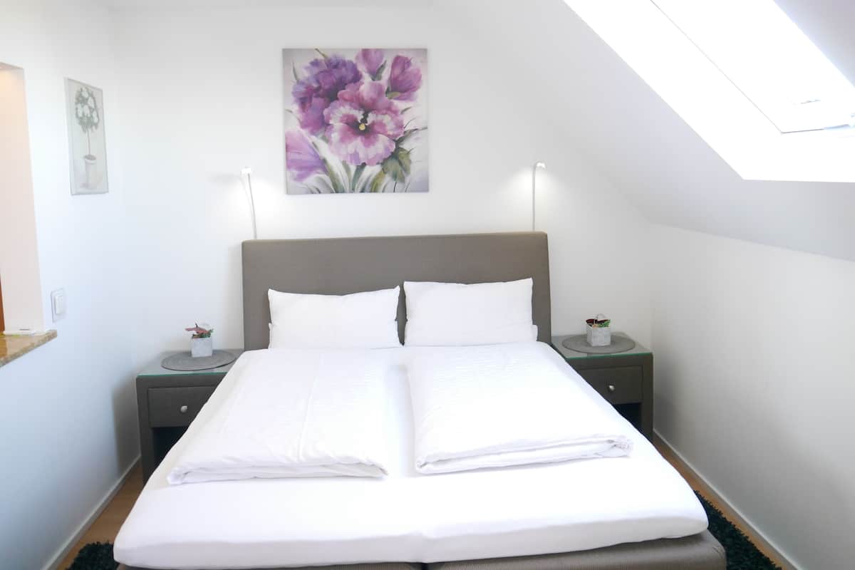 Un lit bien fait dans une pièce dotée d’une lucarne, un tableau représentant des fleurs accroché au mur et une petite table avec une lampe.