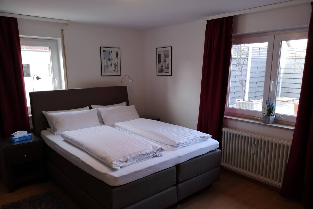 Un lit double bien fait, avec une tête de lit, une table de chevet avec une lampe, une fenêtre avec des rideaux et un radiateur.