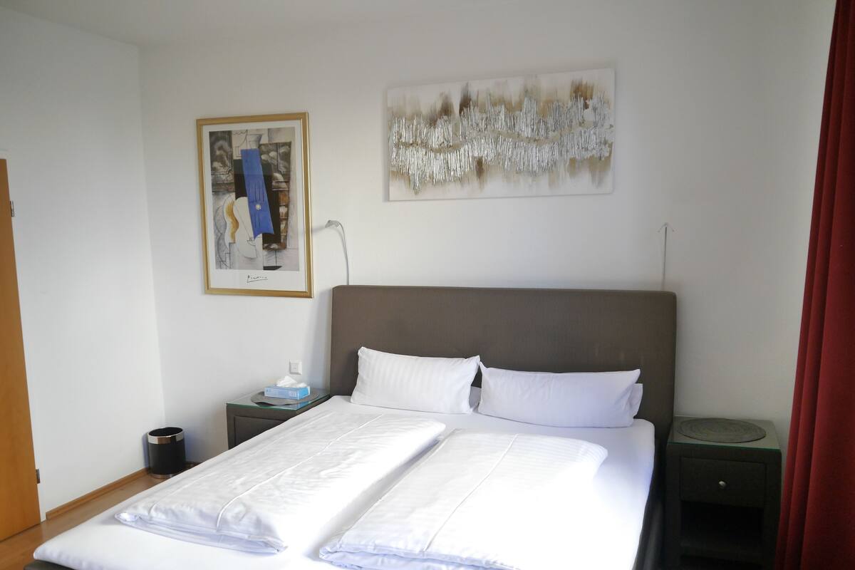 Un lit bien fait, avec du linge de lit blanc et des oreillers, une table de chevet avec une lampe et un tableau encadré au mur.