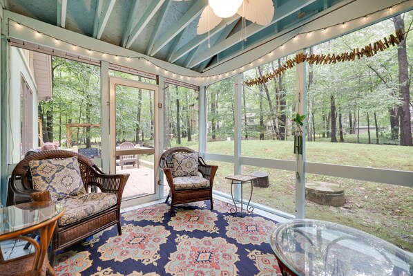 A covered patio area with wicker furniture, a patterned rug, and a view of a wooded area.
