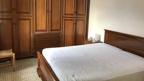 Un dormitorio con armario de madera, cama con ropa blanca, mesita de noche con reloj y suelo estampado.