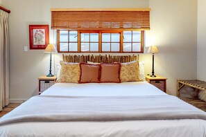 A neatly made bed with a headboard, two bedside lamps, a window with blinds, and a framed picture on the wall. - Somerset Oaks (Cape Town)