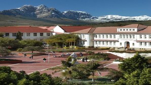 A building with a red-tiled roof and white walls, surrounded by a landscaped area with palm trees and a clear view of snow-capped mountains in the background. - Somerset Oaks (Cape Town)