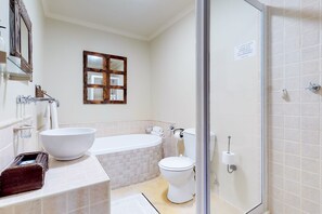 A bathroom with a large white bathtub, a white toilet, and a white sink with a large bowl. There is a mirror above the sink and a window with a wooden frame. - Somerset Oaks (Cape Town)