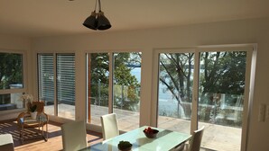 A modern dining area with a glass table, white chairs, and a bowl of fruit. Large windows offer a view of the outdoors. - Direct lake with large south-west balcony (new building 2019) (Überlingen)